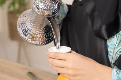 العادات والتقاليد الأصيلة في تقديم القهوة العربية، تصوير مقرب ليد امرأة عربية خليجية اماراتية محجبة ترتدي عباءة سوداء قوم بصب القهوة العربية الساخنة من الدلة الفضية، الاستمتاع لابشرب المشروبات الساخنة