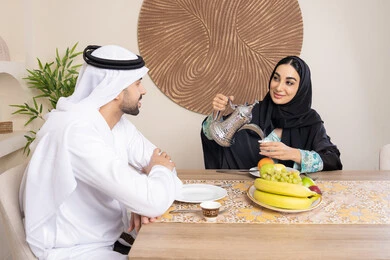 تناول الفواكه الصحية الطازجة، زوجان عربيان خليجيان اماراتيان يجلسان خلف الطاولة في البيت يقومان بشرب القهوة العربية الساخنة بإيماءات السعادة والسرور، مفهوة المحبة والتفام بين الزوجين، جلسة هادئة في المنزل، سيدة محجبة ترتدي العباءة تقوم بصب فنجان من القهوة