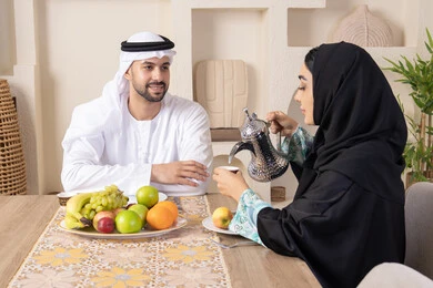 تناول الفواكه الصحية الطازجة، زوجان عربيان خليجيان اماراتيان يجلسان خلف الطاولة في البيت يقومان بشرب القهوة العربية الساخنة بإيماءات السعادة والسرور، مفهوة المحبة والتفام بين الزوجين، جلسة هادئة في المنزل، سيدة محجبة ترتدي العباءة تقوم بصب فنجان من القهوة