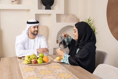 جلسة هادئة في المنزل، تناول الفواكه الصحية الطازجة، زوجان عربيان خليجيان اماراتيان يجلسان خلف الطاولة في البيت يقومان بشرب القهوة العربية الساخنة بإيماءات السعادة والسرور، مفهوة المحبة والتفام بين الزوجين، سيدة محجبة ترتدي العباءة تقوم بصب فنجان من القهوة