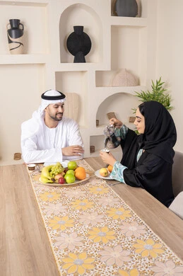 جلسة هادئة في المنزل، تناول الفواكه الصحية الطازجة، زوجان عربيان خليجيان اماراتيان يجلسان خلف الطاولة في البيت يقومان بشرب القهوة العربية الساخنة بإيماءات السعادة والسرور، مفهوة المحبة والتفام بين الزوجين، سيدة محجبة ترتدي العباءة تقوم بصب فنجان من القهوة