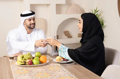 قضاء وقت عائلي ممتع، تناول الفواكه الصحية الطازجة، زوجان عربيان خليجيان اماراتيان يجلسان خلف الطاولة في البيت بإيماءات السعادة والسرور، مفهوم المحبة والتفام بين الزوجين، سيدة محجبة ترتدي العباءة تقوم بتقديم فنجان من القهوة لزوجها