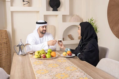 قضاء وقت عائلي ممتع، تناول الفواكه الصحية الطازجة، زوجان عربيان خليجيان اماراتيان يجلسان خلف الطاولة في البيت بإيماءات السعادة والسرور، مفهوم المحبة والتفام بين الزوجين، سيدة محجبة ترتدي العباءة تقوم بتقديم فنجان من القهوة لزوجها