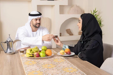 اجواء الأسرة السعيدة، قضاء وقت عائلي ممتع، تناول الفواكه الصحية الطازجة، زوجان عربيان خليجيان اماراتيان يجلسان خلف الطاولة في البيت بإيماءات السعادة والسرور، مفهوم المحبة والتفام بين الزوجين، سيدة محجبة ترتدي العباءة تقوم بتقديم فنجان من القهوة لزوجها