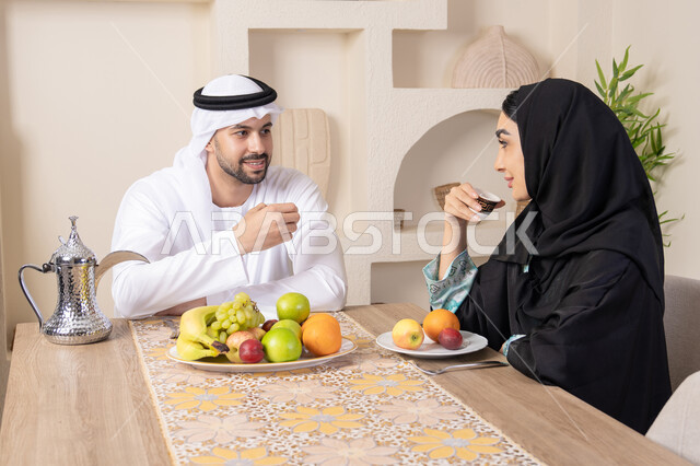 التفاهم والمحبة بين الزوجين، اجواء الأسرة السعيدة، قضاء وقت عائلي ممتع، تناول الفواكه الصحية الطازجة، زوجان عربيان خليجيان اماراتيان يجلسان خلف الطاولة في المنزل يقومان بشرب القهوة العربية الساخنة بإيماءات السعادة والسرور