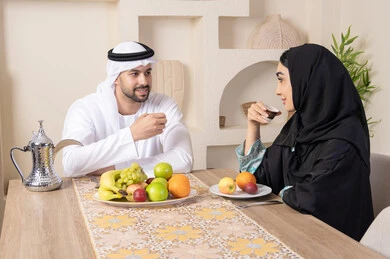التفاهم والمحبة بين الزوجين، اجواء الأسرة السعيدة، قضاء وقت عائلي ممتع، تناول الفواكه الصحية الطازجة، زوجان عربيان خليجيان اماراتيان يجلسان خلف الطاولة في المنزل يقومان بشرب القهوة العربية الساخنة بإيماءات السعادة والسرور