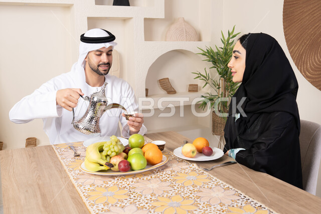 The concept of love and understanding between spouses, a happy family atmosphere, spending enjoyable family time, eating fresh healthy fruits, an Emirati Gulf Arab couple sitting behind the table at home with gestures of happiness and pleasure, a man wearing a kandura and ghutra pouring a cup of coffee for his wife