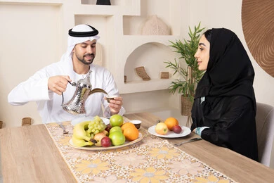مفهوم المحبة والتفاهم بين الزوجين، اجواء الأسرة السعيدة، قضاء وقت عائلي ممتع، تناول الفواكه الصحية الطازجة، زوجان عربيان خليجيان اماراتيان يجلسان خلف الطاولة في البيت بإيماءات السعادة والسرور، رجل يرتدي الكندورة والغترة يقوم بصب فنجان من القهوة لزوجته
