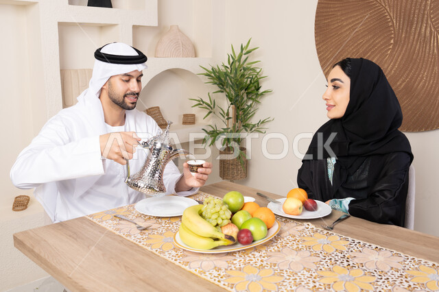 المحبة والتفاهم بين الزوجين، اجواء الأسرة السعيدة، قضاء وقت عائلي ممتع، تناول الفواكه الصحية الطازجة، زوجان عربيان خليجيان اماراتيان يجلسان خلف الطاولة في البيت بإيماءات السعادة والسرور، رجل يرتدي الكندورة والغترة يقوم بصب فنجان من القهوة لزوجته