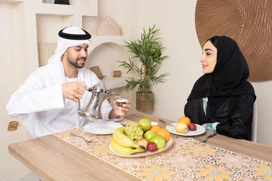 المحبة والتفاهم بين الزوجين، اجواء الأسرة السعيدة، قضاء وقت عائلي ممتع، تناول الفواكه الصحية الطازجة، زوجان عربيان خليجيان اماراتيان يجلسان خلف الطاولة في البيت بإيماءات السعادة والسرور، رجل يرتدي الكندورة والغترة يقوم بصب فنجان من القهوة لزوجته