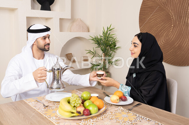 The concept of love and understanding between spouses, a happy family atmosphere, spending enjoyable family time, eating fresh healthy fruits, an Emirati Gulf Arab couple sitting behind the table at home with gestures of happiness and pleasure, a man wearing a kandura and ghutra pouring a cup of coffee for his wife