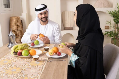 موسم الفاكهة الصحية اللذيذة، التفاهم والمحبة بين الزوجين، اجواء الأسرة السعيدة، قضاء وقت عائلي ممتع، زوجان عربيان خليجيان اماراتيان يجلسان خلف الطاولة في المنزل يقومان بتناول الفواكه الطازجة بإيماءات السعادة والسرور، سناك خفيف ومنعش