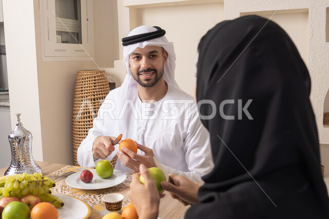 قضاء وقت عائلي ممتع، سناك خفيف ومنعش، موسم الفاكهة الصحية اللذيذة، التفاهم والمحبة بين الزوجين، اجواء الأسرة السعيدة، زوجان عربيان خليجيان اماراتيان يجلسان خلف الطاولة في المنزل يقومان بتناول الفواكه الطازجة بإيماءات السعادة والسرور