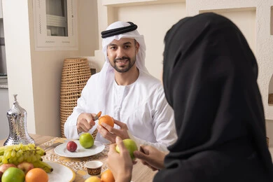 قضاء وقت عائلي ممتع، سناك خفيف ومنعش، موسم الفاكهة الصحية اللذيذة، التفاهم والمحبة بين الزوجين، اجواء الأسرة السعيدة، زوجان عربيان خليجيان اماراتيان يجلسان خلف الطاولة في المنزل يقومان بتناول الفواكه الطازجة بإيماءات السعادة والسرور