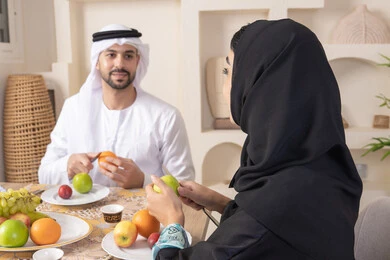 سناك خفيف ومنعش، موسم الفاكهة الصحية اللذيذة، التفاهم والمحبة بين الزوجين، اجواء الأسرة السعيدة، قضاء وقت عائلي ممتع، زوجان عربيان خليجيان اماراتيان يجلسان خلف الطاولة في المنزل يقومان بتناول الفواكه الطازجة بإيماءات السعادة والسرور