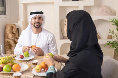 سناك خفيف ومنعش، موسم الفاكهة الصحية اللذيذة، التفاهم والمحبة بين الزوجين، اجواء الأسرة السعيدة، قضاء وقت عائلي ممتع، زوجان عربيان خليجيان اماراتيان يجلسان خلف الطاولة في المنزل يقومان بتناول الفواكه الطازجة بإيماءات السعادة والسرور