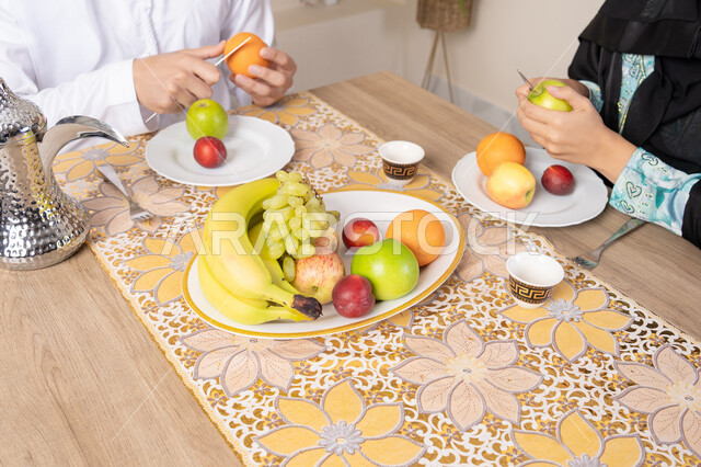 Spending fun family time, light and refreshing snack, the season of delicious healthy fruits, understanding and love between spouses, happy family atmosphere, an Arab Gulf Emirati couple sitting behind the table at home eating fresh fruits with gestures of happiness and pleasure