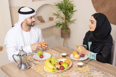 The season of delicious healthy fruits, understanding and love between spouses, a happy family atmosphere, spending enjoyable family time, an Arab Gulf Emirati couple sitting behind the table at home eating fresh fruits with gestures of happiness and pleasure, a light and refreshing snack