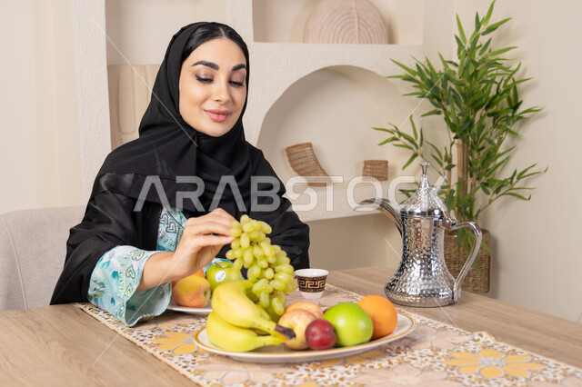 سناك صحي وخفيف، طبق من الفاكهه الطازجة، حسن إكرام الضيف، العادات والتقاليد الأصيلة في تقديم القهوة العربية، امرأة عربية خليجية اماراتية محجبة مبتسمة ترتدي عباءة سوداء تجلس خلف الطاولة وتقوم بأكل الفواكه المنعشة، نمط حياة صحي