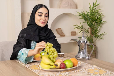 A healthy and light snack, a plate of fresh fruit, hospitality, authentic customs and traditions in serving Arabic coffee, a smiling veiled Emirati Gulf Arab woman wearing a black abaya sitting behind the table and eating refreshing fruit, a healthy lifestyle