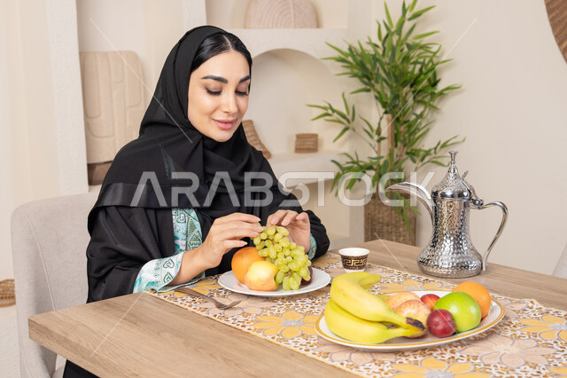 Healthy lifestyle, healthy and light snack, fresh fruit platter, hospitality, authentic customs and traditions of serving Arabic coffee, a smiling veiled Emirati Gulf Arab woman wearing a black abaya sitting behind the table and eating refreshing fruits