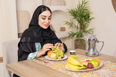 Healthy lifestyle, healthy and light snack, fresh fruit platter, hospitality, authentic customs and traditions of serving Arabic coffee, a smiling veiled Emirati Gulf Arab woman wearing a black abaya sitting behind the table and eating refreshing fruits