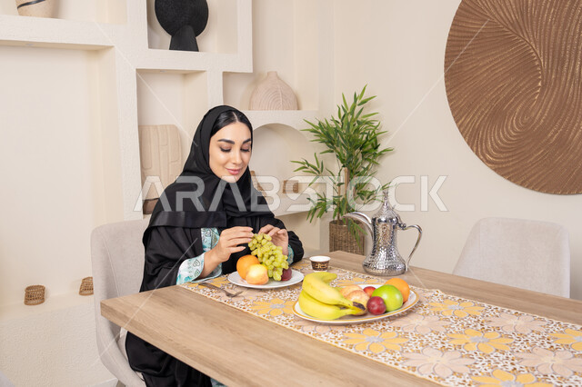 سناك صحي وخفيف، طبق من الفاكهه الطازجة، حسن إكرام الضيف، العادات والتقاليد الأصيلة في تقديم القهوة العربية، امرأة عربية خليجية اماراتية محجبة مبتسمة ترتدي عباءة سوداء تجلس خلف الطاولة وتقوم بأكل الفواكه المنعشة، نمط حياة صحي