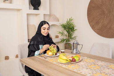 A healthy and light snack, a plate of fresh fruit, hospitality, authentic customs and traditions in serving Arabic coffee, a smiling veiled Emirati Gulf Arab woman wearing a black abaya sitting behind the table and eating refreshing fruit, a healthy lifestyle