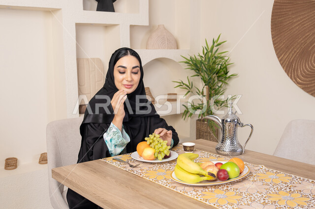 A healthy and light snack, a plate of fresh fruit, hospitality, authentic customs and traditions in serving Arabic coffee, a smiling veiled Emirati Gulf Arab woman wearing a black abaya sitting behind the table and eating refreshing fruit, a healthy lifestyle