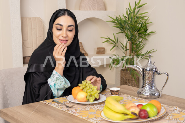 Healthy lifestyle, healthy and light snack, fresh fruit platter, hospitality, authentic customs and traditions of serving Arabic coffee, a smiling veiled Emirati Gulf Arab woman wearing a black abaya sitting behind the table and eating refreshing fruits
