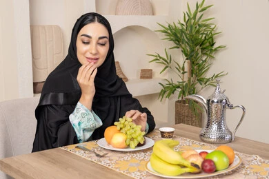Healthy lifestyle, healthy and light snack, fresh fruit platter, hospitality, authentic customs and traditions of serving Arabic coffee, a smiling veiled Emirati Gulf Arab woman wearing a black abaya sitting behind the table and eating refreshing fruits