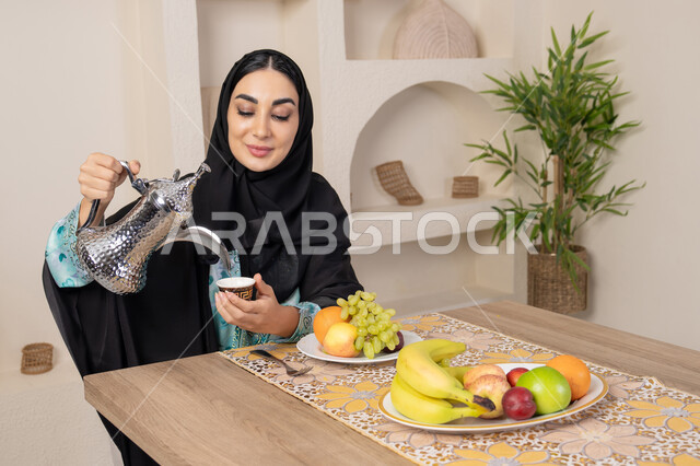 طبق من الفاكهه الطازجة، حسن إكرام الضيف، العادات والتقاليد الأصيلة في تقديم القهوة العربية، امرأة عربية خليجية اماراتية محجبة مبتسمة ترتدي عباءة سوداء تجلس خلف الطاولة وتقوم بصب القهوة العربية الساخنة من الدلة الفضية، الاستمتاع بشرب المشروبات الساخنة