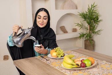 A plate of fresh fruit, hospitality, authentic customs and traditions of serving Arabic coffee, a smiling veiled Emirati Gulf Arab woman wearing a black abaya sitting behind the table pouring hot Arabic coffee from a silver coffee pot, enjoying drinking hot drinks