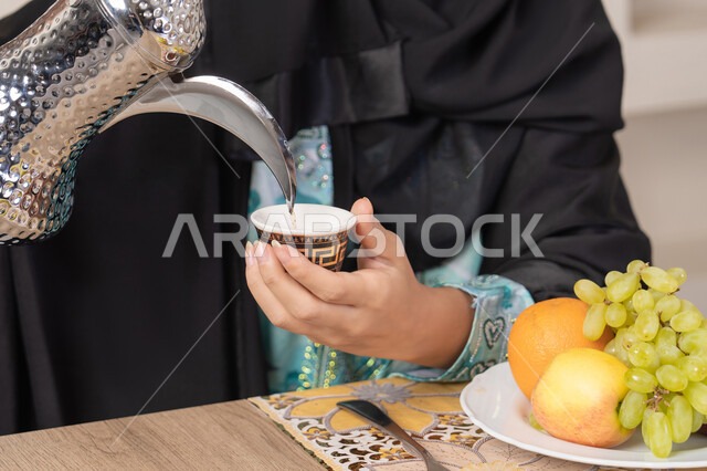 حسن إكرام الضيف، العادات والتقاليد الأصيلة في تقديم القهوة العربية، تصوير مقرب ليد امرأة عربية خليجية اماراتية محجبة ترتدي عباءة سوداء قوم بصب القهوة العربية الساخنة من الدلة الفضية، الاستمتاع لابشرب المشروبات الساخنة، طبق من الفاكهه الطازجة
