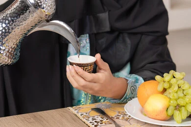 Good hospitality, authentic customs and traditions in serving Arabic coffee, close-up of the hand of a veiled Emirati Gulf Arab woman wearing a black abaya pouring hot Arabic coffee from a silver coffee pot, enjoying a hot beverage, a plate of fresh fruit