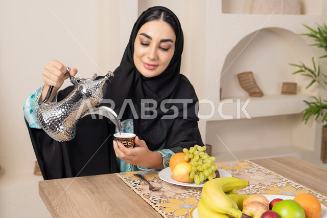 طبق من الفاكهه الطازجة، حسن إكرام الضيف، العادات والتقاليد الأصيلة في تقديم القهوة العربية، امرأة عربية خليجية اماراتية محجبة مبتسمة ترتدي عباءة سوداء تجلس خلف الطاولة وتقوم بصب القهوة العربية الساخنة من الدلة الفضية، الاستمتاع بشرب المشروبات الساخنة