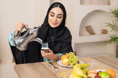 طبق من الفاكهه الطازجة، حسن إكرام الضيف، العادات والتقاليد الأصيلة في تقديم القهوة العربية، امرأة عربية خليجية اماراتية محجبة مبتسمة ترتدي عباءة سوداء تجلس خلف الطاولة وتقوم بصب القهوة العربية الساخنة من الدلة الفضية، الاستمتاع بشرب المشروبات الساخنة