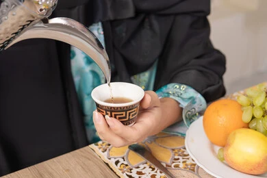 Good hospitality, authentic customs and traditions in serving Arabic coffee, close-up of the hand of a veiled Emirati Gulf Arab woman wearing a black abaya pouring hot Arabic coffee from a silver coffee pot, enjoying a hot beverage, a plate of fresh fruit