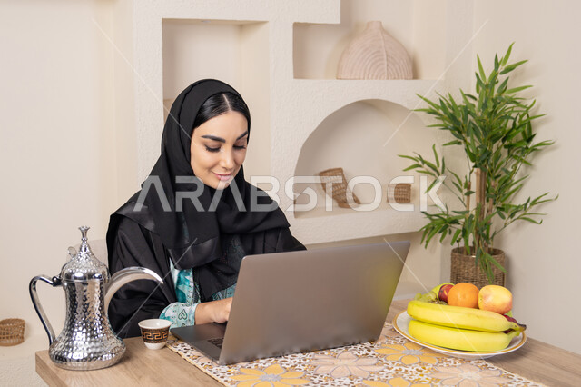 جلسة هادئة في املنزل، سيدة إماراتية يقوم بأداء مهام العمل عن بعد، الاستمتاع بشرب القهوة العربية، دمج التكنولوجيا الحديثة مع الروتين اليومي، امرأة عربية خليجية سعودية ترتدي العباءة والحجاب تجلس خلف طاولة في المنزل تستخدم الحاسوب المحمول، أنواع الفاكهة الطازجة