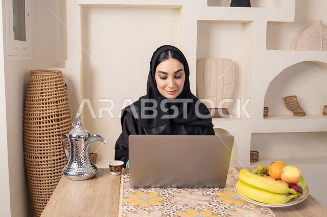 دمج التكنولوجيا الحديثة مع الروتين اليومي، تعابير الاندماج والتركيز، سيدة إماراتية يقوم بأداء مهام العمل عن بعد، الاستمتاع بشرب القهوة العربية، امرأة عربية خليجية سعودية ترتدي العباءة والحجاب تجلس خلف طاولة في المنزل تستخدم الحاسوب المحمول، أنواع الفاكهة الطازجة