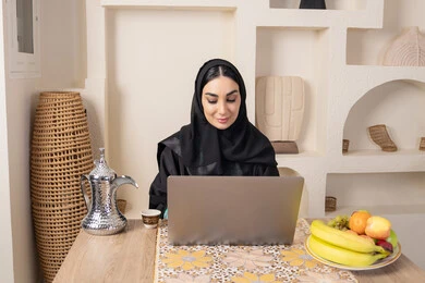 Integrating modern technology with daily routine, expressions of integration and focus, Emirati woman performing remote work tasks, enjoying drinking Arabic coffee, Saudi Arabian Gulf woman wearing abaya and hijab sitting behind a table at home using a laptop, types of fresh fruits