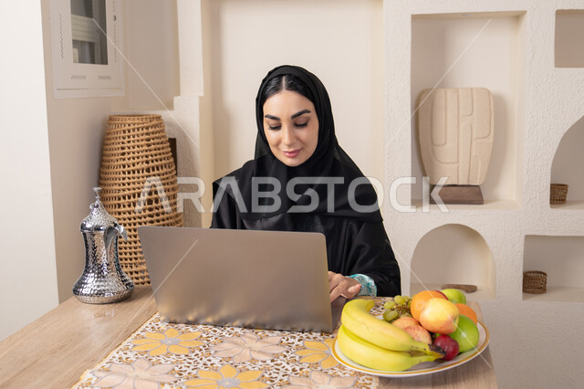 تعابير الاندماج والتركيز، سيدة إماراتية يقوم بأداء مهام العمل عن بعد، الاستمتاع بشرب القهوة العربية، دمج التكنولوجيا الحديثة مع الروتين اليومي، امرأة عربية خليجية سعودية ترتدي العباءة والحجاب تجلس خلف طاولة في المنزل تستخدم الحاسوب المحمول، أنواع الفاكهة الطازجة
