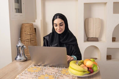 تعابير الاندماج والتركيز، سيدة إماراتية يقوم بأداء مهام العمل عن بعد، الاستمتاع بشرب القهوة العربية، دمج التكنولوجيا الحديثة مع الروتين اليومي، امرأة عربية خليجية سعودية ترتدي العباءة والحجاب تجلس خلف طاولة في المنزل تستخدم الحاسوب المحمول، أنواع الفاكهة الطازجة