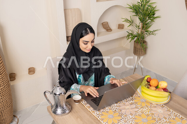 تعابير الاندماج والتركيز، سيدة إماراتية يقوم بأداء مهام العمل عن بعد، الاستمتاع بشرب القهوة العربية، دمج التكنولوجيا الحديثة مع الروتين اليومي، امرأة عربية خليجية سعودية ترتدي العباءة والحجاب تجلس خلف طاولة في المنزل تستخدم الحاسوب المحمول، أنواع الفاكهة الطازجة