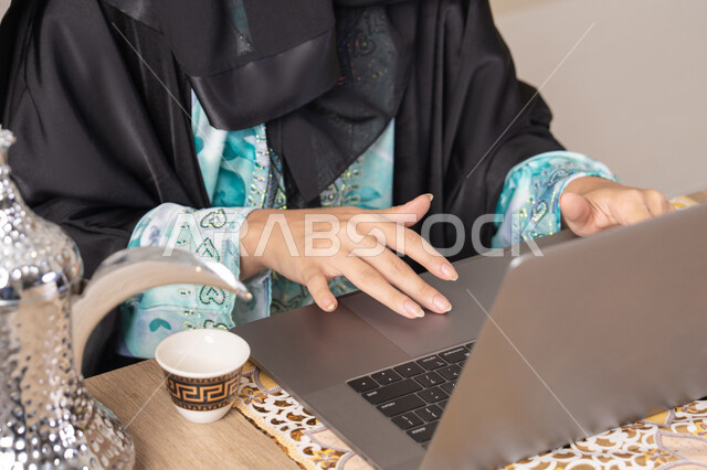 التقنيات الحديثة والمتطورة، تعابير الاندماج والتركيز، سيدة إماراتية يقوم بأداء مهام العمل عن بعد، الاستمتاع بشرب القهوة العربية، تصوير مقرب امرأة عربية خليجية سعودية ترتدي العباءة تجلس خلف طاولة في المنزل تستخدم الحاسوب المحمول، أنواع الفاكهة الطازجة