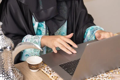 Modern and advanced technologies, expressions of integration and focus, Emirati woman performing remote work tasks, enjoying drinking Arabic coffee, close-up of a Saudi Arabian Gulf woman wearing an abaya sitting behind a table at home using a laptop, types of fresh fruits