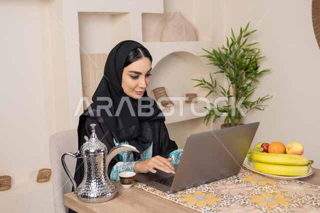 جلسة هادئة في املنزل، سيدة إماراتية يقوم بأداء مهام العمل عن بعد، الاستمتاع بشرب القهوة العربية، دمج التكنولوجيا الحديثة مع الروتين اليومي، امرأة عربية خليجية سعودية ترتدي العباءة والحجاب تجلس خلف طاولة في المنزل تستخدم الحاسوب المحمول، أنواع الفاكهة الطازجة