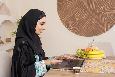 الفاكهه الطازجة والمنعشة، تعابير الاندماج والتركيز، سيدة إماراتية تقوم بأداء مهام العمل عن بعد، الاستمتاع بشرب القهوة العربية، تصوير من الجانب امرأة عربية خليجية سعودية ترتدي العباءة والحجاب تجلس خلف طاولة في المنزل تستخدم الحاسوب المحمول
