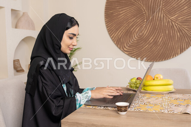 الفاكهه الطازجة والمنعشة، تعابير الاندماج والتركيز، سيدة إماراتية تقوم بأداء مهام العمل عن بعد، الاستمتاع بشرب القهوة العربية، تصوير من الجانب امرأة عربية خليجية سعودية ترتدي العباءة والحجاب تجلس خلف طاولة في المنزل تستخدم الحاسوب المحمول