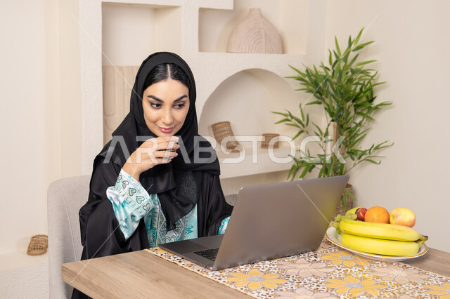 جلسة هادئة في املنزل، سيدة إماراتية يقوم بأداء مهام العمل عن بعد، الاستمتاع بشرب القهوة العربية، دمج التكنولوجيا الحديثة مع الروتين اليومي، امرأة عربية خليجية سعودية ترتدي العباءة والحجاب تجلس خلف طاولة في المنزل تستخدم الحاسوب المحمول، أنواع الفاكهة الطازجة